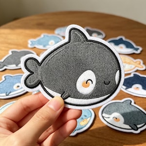 May include: A circular embroidered patch featuring a stylized, cartoon-like orca whale with a dark gray body, a white belly, and a white eye patch with a small orange cheek detail. The patch has a thick black border and a white outer edge.