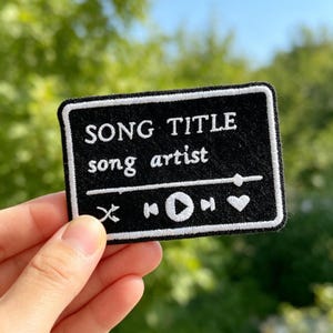 May include: A rectangular black embroidered patch featuring white stitching that mimics a music player interface. The text reads SONG TITLE and song artist, accompanied by a progress bar, shuffle, skip, play, pause, and heart icons.