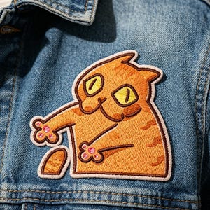 May include: An embroidered patch featuring an orange tabby cat with wide yellow eyes and pink paw pads, stitched onto a blue denim jacket. The cat is depicted in a reaching pose with a dark brown outline, creating a textured fabric detail.
