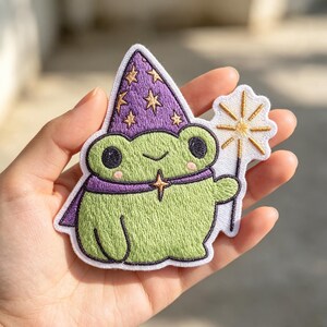 May include: An embroidered patch featuring a light green frog wearing a purple wizard hat decorated with gold stars. The frog has a purple cape, pink cheeks, and holds a gold sparkler wand. The patch has a white border and stitched details.