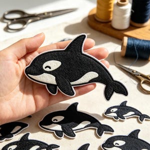 May include: An embroidered iron-on patch shaped like an orca whale. The patch features a black body with a white underbelly and a white eye patch, outlined in white thread. The design depicts a stylized orca with a curved dorsal fin and tail.