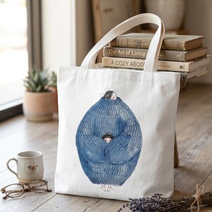 Könnte beinhalten: Weiße Canvas-Tragetasche mit einer Aquarellillustration einer Person und eines Kindes, die in einem großen blauen Pullover eingehüllt sind. Die Tasche hat weiße Griffe und steht auf einer Holzoberfläche. Eine kleine Tasse und eine Brille befinden sich in der Nähe.