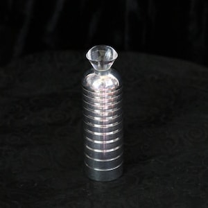 May include: A silver-colored perfume bottle with a clear, diamond-shaped stopper. The bottle has a cylindrical body with horizontal ridges. The background is dark.