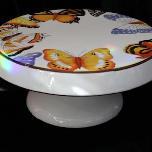 Puede incluir: Un soporte para pasteles de cerámica blanca con una parte superior circular decorada con un patrón colorido de varias mariposas en tonos naranja, amarillo y azul. El soporte tiene un acabado liso y brillante y una base de pedestal ancha.