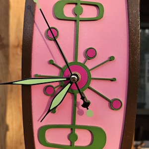 May include: A mid-century modern wall clock with a pink face and a brown wooden frame. The clock features green and pink geometric designs and black clock hands with green tips. The clock measures approximately 12 inches in height.