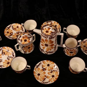 May include: A vintage tea set featuring a white background with brown and orange floral designs. The set includes a teapot, sugar bowl, cups, and saucers. The tea set is arranged on a dark surface.