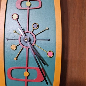 May include: A mid-century modern wall clock with a teal background and a yellow and orange frame. The clock features a red and yellow atomic design with black clock hands. The clock's design is reminiscent of the atomic age.