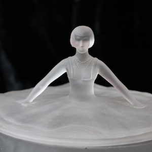 May include: A frosted glass figurine of a ballerina with arms outstretched, wearing a dress. The sculpture is set against a dark background, highlighting the delicate details of the dancer's form and attire.
