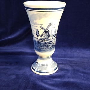 May include: A white and blue ceramic vase with a Dutch windmill scene. The vase has a flared top, a pedestal base, and is decorated with a blue and white landscape design. The vase is approximately 8 inches tall.