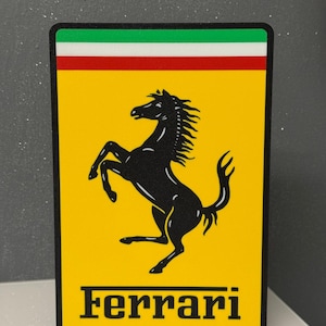 Op de afbeelding: Een rechthoekig bord met een gele achtergrond en een zwart silhouet van een steigerend paard in het midden. Boven het paard bevindt zich een horizontale groen-wit-rode streep. Het woord Ferrari staat in zwarte letters onderaan het bord.