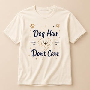 May include: A cream-colored short-sleeve t-shirt featuring the dark blue text "Dog Hair, Don't Care" centered on the chest. The design includes a gold-colored line art illustration of a dog face, a heart, paw prints, and small star accents.