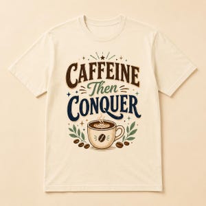 May include: A cream-colored short-sleeve t-shirt featuring a graphic design with the text CAFFEINE Then CONQUER in brown and dark blue lettering. Below the text is a coffee cup illustration with steam, surrounded by green leaves and coffee beans.