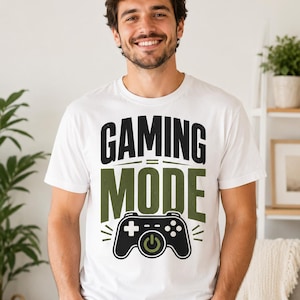 May include: A white short-sleeved t-shirt featuring the black and olive green text GAMING MODE and a graphic of a black game controller with an olive green power button symbol centered below the text on the front of the garment.