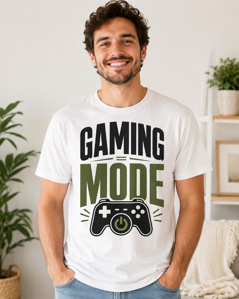 May include: A white short-sleeved t-shirt featuring the black and olive green text GAMING MODE and a graphic of a black game controller with an olive green power button symbol centered below the text on the front of the garment.