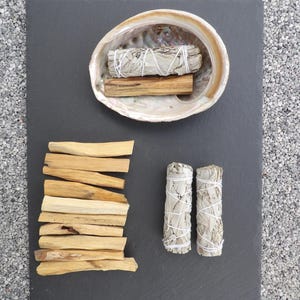 Peut inclure: Une composition d'articles spirituels disposés sur une ardoise gris foncé. L'arrangement comprend des fagots de sauge blanche liés avec de la ficelle blanche, des bâtons de palo santo et une coquille d'ormeau décorative.