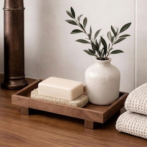 Può includere: Un vassoio rettangolare in legno scuro con quattro piedini sostiene una saponetta rettangolare color bianco sporco appoggiata su una luffa beige testurizzata, accanto a un piccolo vaso in ceramica maculata color bianco sporco con un ramo verde.