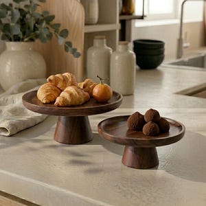 May include: Two dark brown wooden pedestal stands with circular tops and tapered bases. The larger stand holds three golden-brown croissants and a small orange fruit, while the smaller stand holds four round dark brown chocolate truffles.