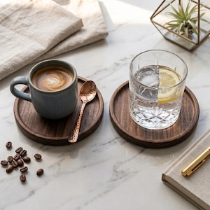 May include: Two round dark wood coasters, one holding a blue ceramic mug filled with coffee and a small hammered copper spoon, the other holding a clear glass tumbler with ice and a lemon slice, accompanied by scattered roasted coffee beans.