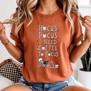 Puede incluir: Una camiseta de manga corta color naranja quemado con texto blanco que dice HOCUS POCUS I NEED COFFEE TO FOCUS. El diseño incluye pequeñas ilustraciones de calabazas, estrellas y un esqueleto acostado leyendo un libro con una taza de café.