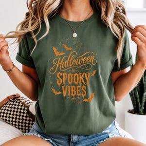 以下が含まれることがあります： オレンジ色のグラフィックプリントが施されたオリーブグリーンのTシャツ。「Halloween SPOOKY VIBES」という文字が、滴るようなスタイリッシュなフォントで描かれています。デザインには、小さなオレンジ色のコウモリのシルエットと、文字を囲む細いクモの巣の線が含まれています。