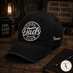 Op de afbeelding: Een zwarte baseballpet met een witte cirkelvormige geborduurde patch op de voorkant met de tekst Cool Dads Club in een gestileerd lettertype. De zijkant van de pet is geborduurd met de naam Thomas in witte schrijfletters.