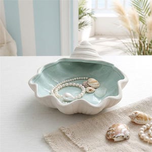 Puede incluir: Un cuenco de cerámica con forma de concha marina con un exterior blanco mate y un interior verde menta claro brillante. Dentro del cuenco hay un collar de perlas blancas y pequeñas conchas naturales. El cuenco tiene una parte superior en espiral.