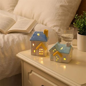 Pode incluir: Dois suportes de cerâmica para velas em forma de casa com paredes brancas, telhados azuis e detalhes amarelos. A casa mais alta apresenta um recorte em forma de estrela, enquanto a menor tem um recorte em forma de lua crescente.