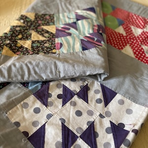 Könnte beinhalten: Eine gefaltete Steppdecke mit Patchwork-Design. Die Steppdecke weist verschiedene Stoffmuster auf, darunter Blumenmuster, Tupfen und geometrische Formen, in Rot-, Lila-, Blau- und Grautönen. Die Ränder sind mit einem einfarbigen grauen Stoff versehen.
