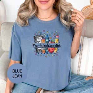 May include: A blue jean colored t-shirt featuring a graphic print of a composition notebook, glue stick, crayon, apple, and pencil with the text Pre-Kindergarten. The design includes colorful stars and is centered on the chest of the garment.