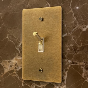 May include: A rectangular light switch plate with a brushed gold finish and a textured surface. The plate features a central toggle switch with a knurled gold handle and a white base, secured by two flat-head screws at the top and bottom.