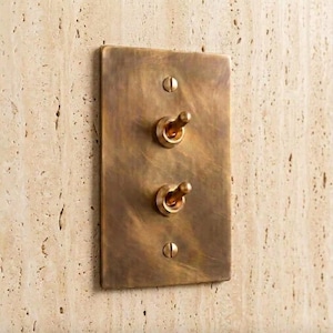 May include: A rectangular brass light switch plate with a brushed, weathered finish. The plate features two vertical toggle switches positioned in the center and is secured to the wall with two matching brass screws at the top and bottom.