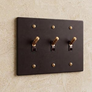 May include: A rectangular matte black triple light switch wall plate featuring three brass toggle switches with a textured knurled finish. The plate is secured to the wall with six matching brass flathead screws positioned at the top and bottom.