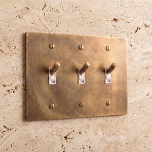 May include: A rectangular triple light switch wall plate made of brushed brass with a weathered finish. The plate features three white toggle switches with textured, knurled brass extensions and is secured to the wall with six matching brass screws.