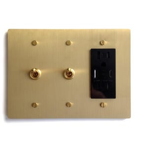 May include: A rectangular brushed brass wall plate featuring two gold-toned metal toggle switches and one black electrical outlet with integrated USB-A and USB-C charging ports. The plate is secured with six matching brass screws.