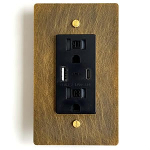 May include: A rectangular electrical wall outlet with a black faceplate featuring two standard power sockets, one USB-A port, and one USB-C port. The outlet is mounted within a brushed brass metal cover plate secured by two flathead brass screws.