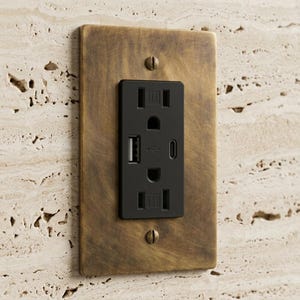 May include: A rectangular electrical wall socket with a matte black finish, featuring two standard power outlets, one USB-A port, and one USB-C port. The socket is mounted within a brushed antique brass metal wall plate secured by two screws.