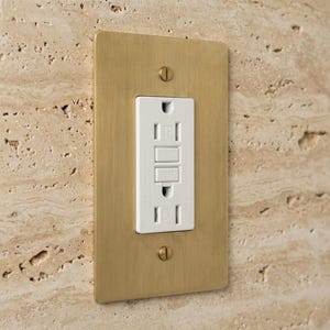 May include: A rectangular wall plate with a brushed brass finish and two matching screws, surrounding a white GFCI electrical outlet. The outlet features a central reset button, a test button, and the text TR printed on the white plastic surface.