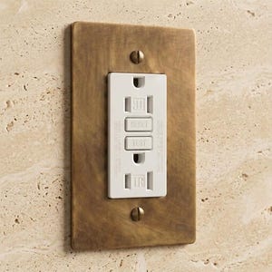 May include: A rectangular electrical GFCI outlet with a white plastic face featuring Reset and Test buttons, secured by two flathead screws into a brushed antique brass metal wall plate with a textured, weathered finish and rounded corners.