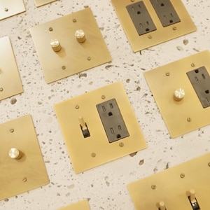 May include: A collection of square brass wall plates featuring various configurations of black electrical outlets, toggle switches, and circular rotary dimmer knobs, all secured with small matching brass screws on a speckled white surface.