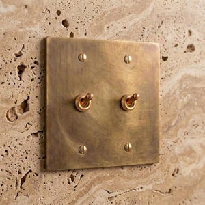 May include: A square, brushed brass double toggle light switch plate featuring two central metal toggles and four matching screws at the corners. The plate has a warm, metallic finish with visible texture and is mounted on a stone wall.