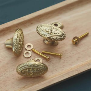 May include: A set of antique brass cabinet knobs and hardware. The knobs have an oval shape with ornate floral detailing. Included are screws and washers for installation. The hardware is displayed on a light wood tray.