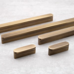Può includere: Una collezione di pomelli e maniglie per mobili in ottone antico. L'hardware presenta un design rettangolare con bordi arrotondati. La finitura ha un aspetto caldo e invecchiato. Le maniglie sono di varie dimensioni, adatte per cassetti e ante.