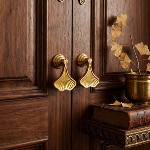 Puede incluir: Primer plano de una puerta de madera con dos manijas de latón. Las manijas tienen forma de hojas de ginkgo estilizadas. La puerta es de color marrón oscuro. Una pequeña maceta de latón con hojas secas está sobre una estantería.