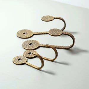 May include: Four antique brass wall hooks with a hammered texture. Each hook has a circular mounting plate with a central hole and a curved hook. The hooks are arranged in a staggered pattern.