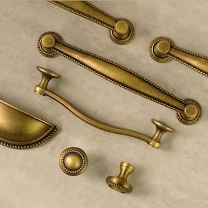 May include: An assortment of antique brass cabinet hardware, including handles and knobs. The hardware features a detailed design with a warm, golden hue, and a beaded border. The collection includes various sizes and shapes for different cabinet styles.