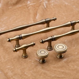 May include: A collection of antique brass cabinet hardware, including long bar pulls and round knobs. The hardware has a vintage aesthetic with a warm, aged finish. The knobs and pulls are designed for furniture and cabinetry.