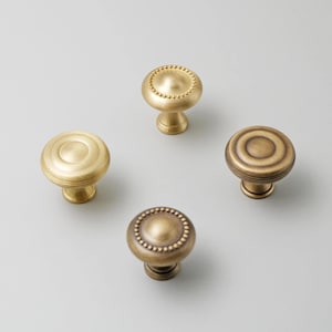 Pode incluir: Quatro puxadores de armário de latão em vários designs. Dois puxadores têm um design circular, um com borda de contas. Os outros dois puxadores têm um design canelado. Todos os puxadores têm uma base redonda e um acabamento dourado.