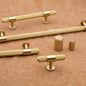 May include: A collection of gold-colored cabinet hardware, including bar pulls and cylindrical knobs. The hardware features a textured, cross-hatched pattern on the handles and knobs. The T-shaped pulls and knobs are displayed on a brown surface.