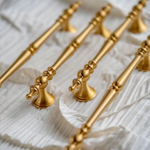 Könnte beinhalten: Mehrere goldfarbene Schrankgriffe sind auf einer weißen, strukturierten Oberfläche angeordnet. Die Griffe haben ein detailliertes Design mit dekorativem Sockel und einem langen, schlanken Griff. Geeignet für Möbel oder Schränke.