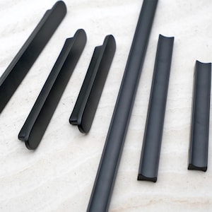 Può includere: Una collezione di maniglie e pomelli per mobili neri opachi di varie dimensioni. L'hardware presenta un design elegante e moderno con un'estetica liscia e minimalista. Le maniglie sono disposte su una superficie chiara.